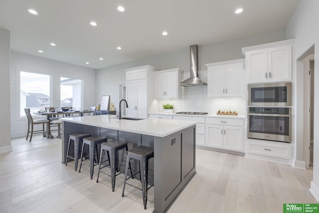 kitchen with stainless steel appliances, light wood-style floors, a kitchen island with sink, a sink, and wall chimney exhaust hood