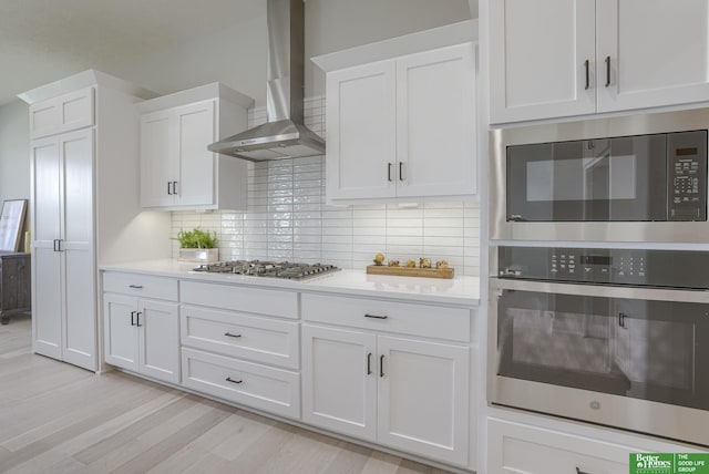 kitchen featuring white cabinets, decorative backsplash, wall chimney exhaust hood, stainless steel appliances, and light countertops