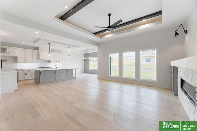 unfurnished living room featuring ceiling fan with notable chandelier, a stone fireplace, a tray ceiling, and light wood-style flooring