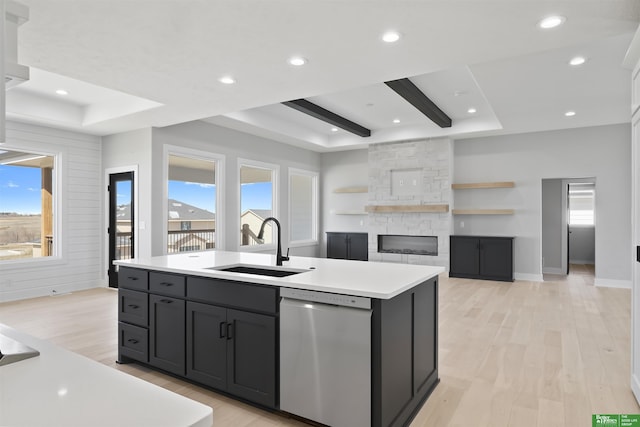 kitchen with a sink, light wood-style floors, stainless steel dishwasher, a raised ceiling, and a center island with sink