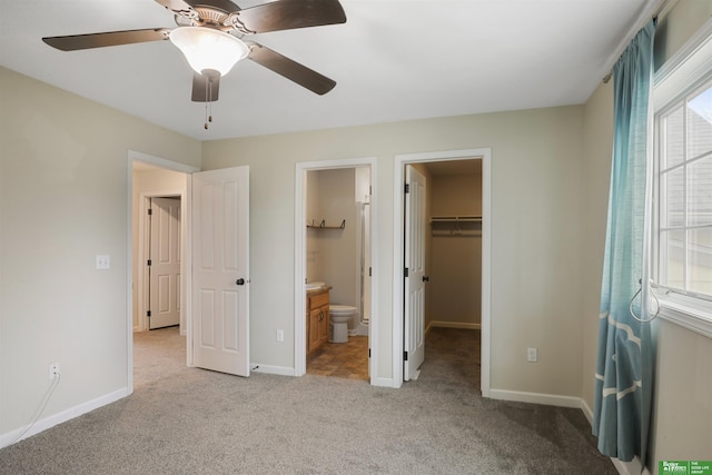 unfurnished bedroom featuring a walk in closet, ensuite bathroom, light carpet, ceiling fan, and baseboards