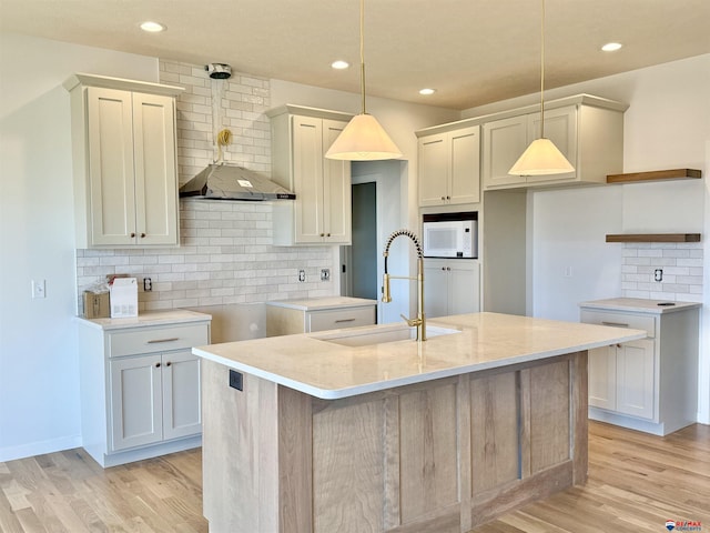 kitchen featuring a center island with sink, light wood finished floors, hanging light fixtures, white microwave, and a sink