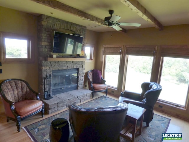 living area with beam ceiling, ceiling fan, a stone fireplace, and wood finished floors