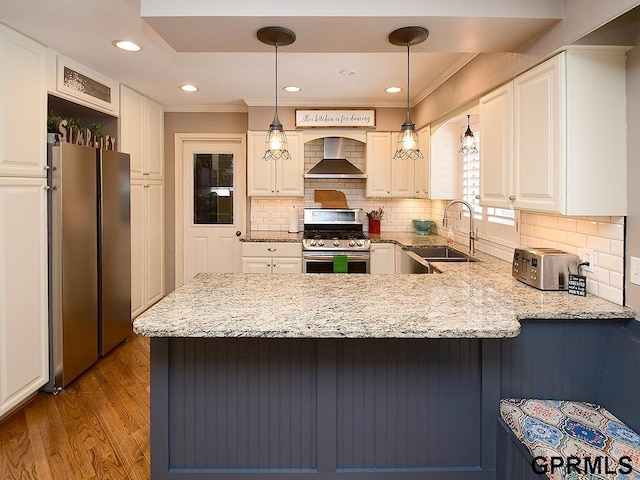 kitchen featuring stainless steel appliances, a sink, wood finished floors, a peninsula, and wall chimney exhaust hood