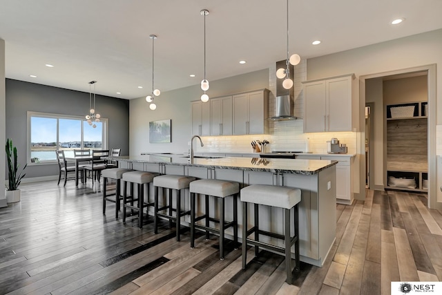 kitchen featuring wood finished floors, a sink, wall chimney exhaust hood, tasteful backsplash, and a center island with sink