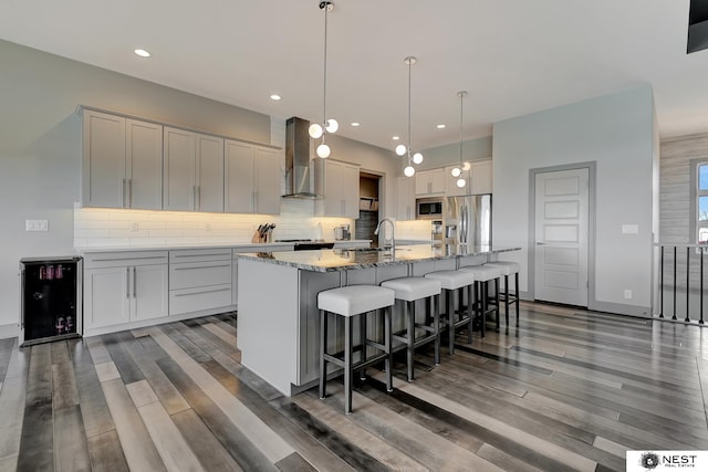 kitchen featuring stainless steel appliances, tasteful backsplash, an island with sink, beverage cooler, and wall chimney exhaust hood