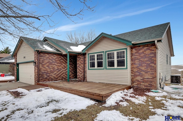 snow covered property with brick siding, roof with shingles, a deck, a garage, and cooling unit