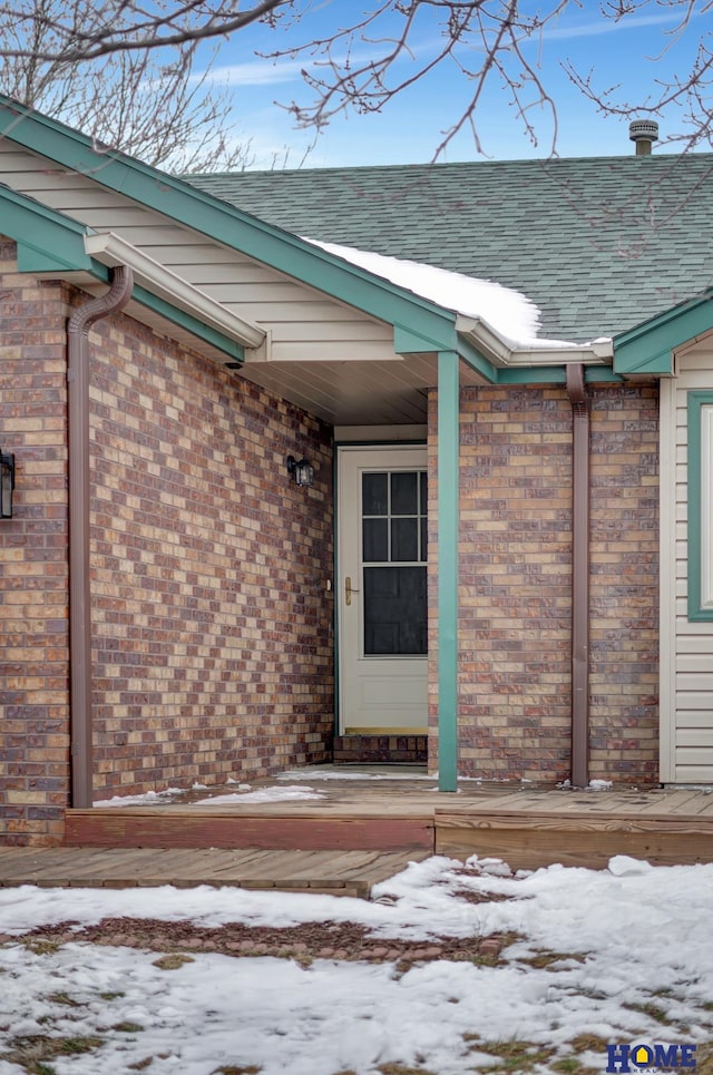 snow covered property entrance featuring brick siding and a shingled roof