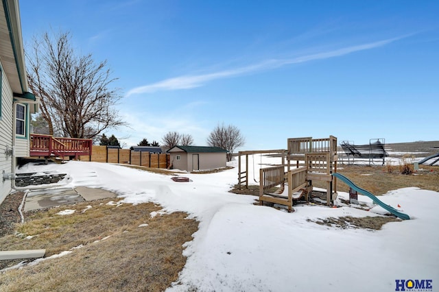 snowy yard featuring a playground, a storage unit, fence, an outdoor structure, and a wooden deck