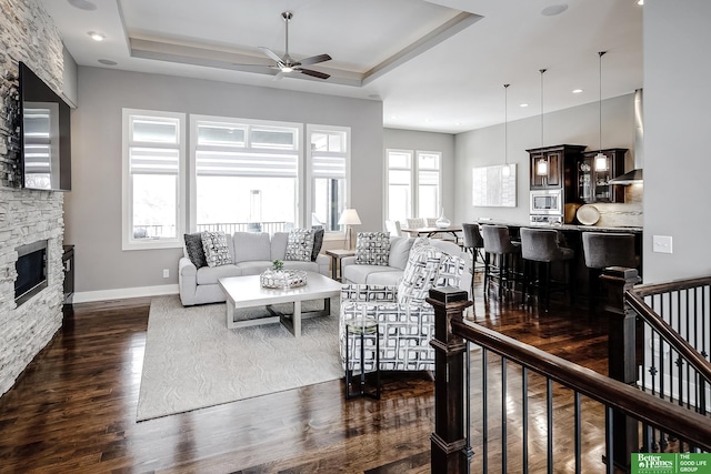 living room featuring a raised ceiling, baseboards, a stone fireplace, and dark wood-style flooring