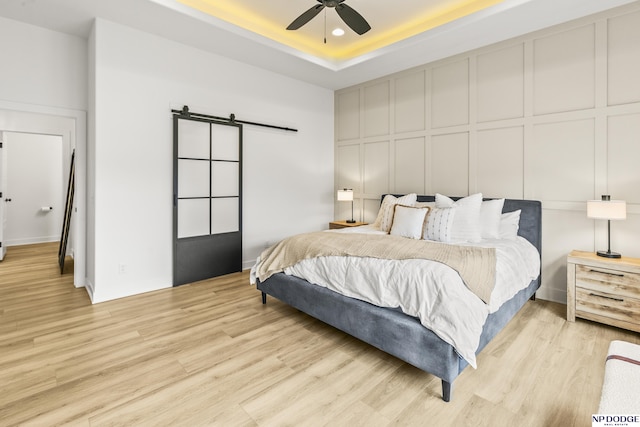 bedroom featuring a barn door, light wood-style flooring, a decorative wall, a ceiling fan, and a tray ceiling