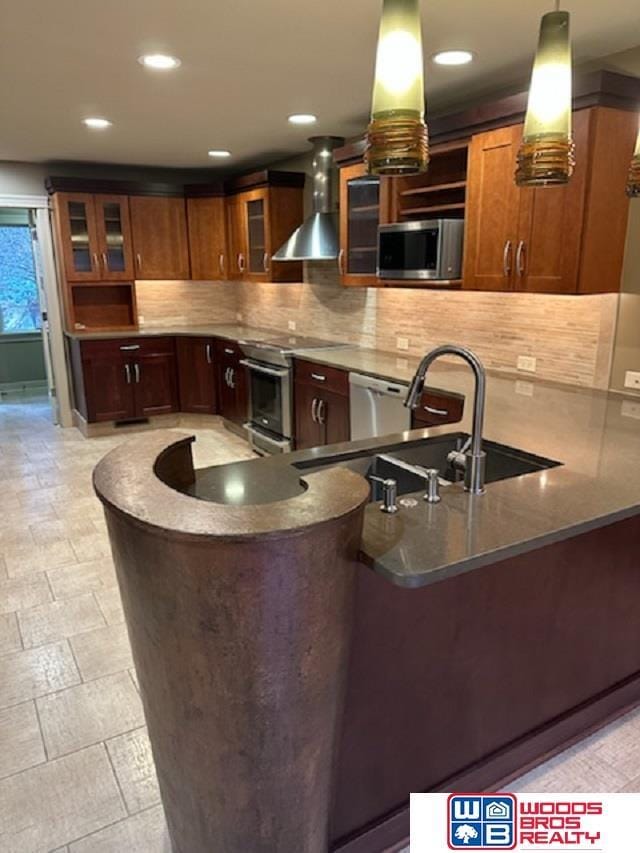 kitchen featuring stainless steel appliances, a peninsula, a sink, backsplash, and wall chimney exhaust hood