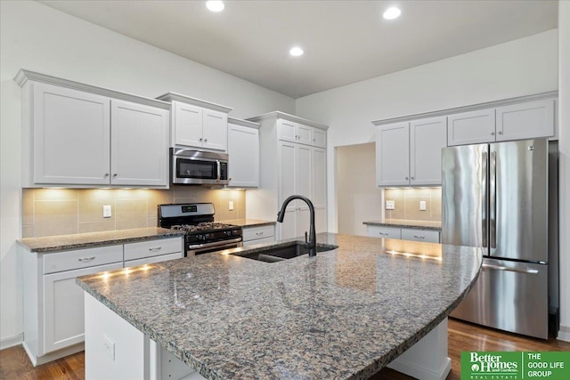 kitchen featuring a kitchen island with sink, stainless steel appliances, a sink, tasteful backsplash, and dark stone countertops