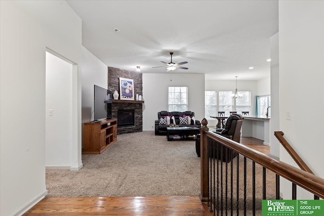 carpeted living area featuring ceiling fan with notable chandelier, baseboards, wood finished floors, and a stone fireplace