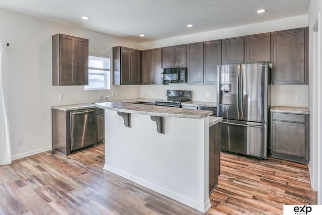 kitchen with dark brown cabinetry, wood finished floors, a kitchen breakfast bar, light countertops, and black appliances