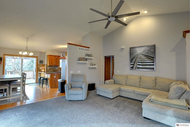 living area with high vaulted ceiling, light carpet, ceiling fan with notable chandelier, visible vents, and light wood-type flooring