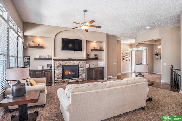 living room featuring baseboards, a textured ceiling, a stone fireplace, and ceiling fan