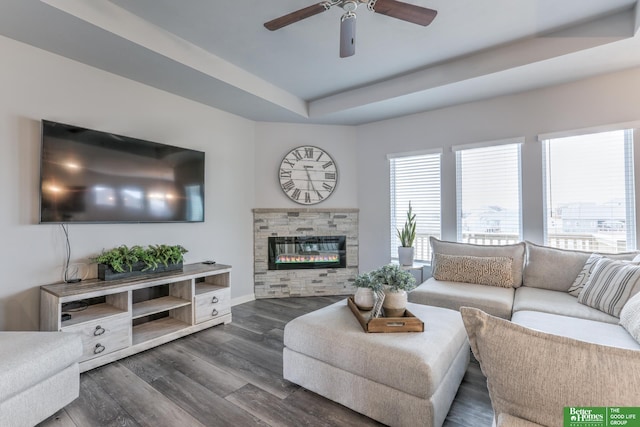 living room with a raised ceiling, dark wood-type flooring, ceiling fan, a stone fireplace, and baseboards