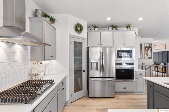 kitchen with stainless steel appliances, light countertops, gray cabinets, wall chimney exhaust hood, and light wood finished floors