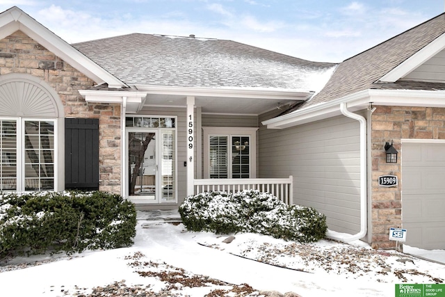 snow covered property entrance featuring a garage, stone siding, a shingled roof, and a porch