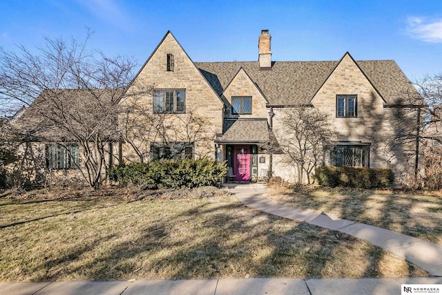 tudor house featuring stone siding, a shingled roof, a chimney, and a front yard