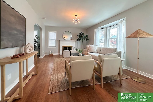 living room featuring baseboards, arched walkways, wood finished floors, an inviting chandelier, and a brick fireplace