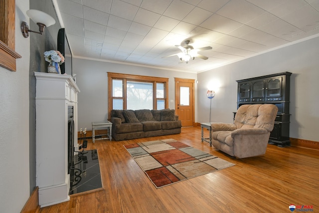 living room featuring crown molding, a fireplace with flush hearth, ceiling fan, wood finished floors, and baseboards