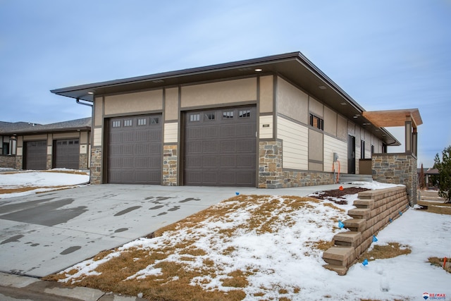 view of snow covered exterior with stone siding, stucco siding, and an attached garage