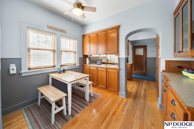 kitchen with brown cabinetry, light wood-style flooring, arched walkways, ceiling fan, and glass insert cabinets