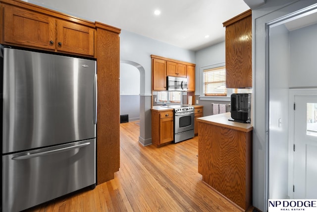 kitchen with brown cabinetry, appliances with stainless steel finishes, light wood-style floors, and arched walkways