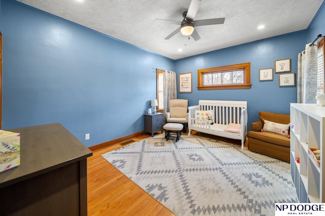 bedroom with wood finished floors, baseboards, ceiling fan, a nursery area, and a textured ceiling