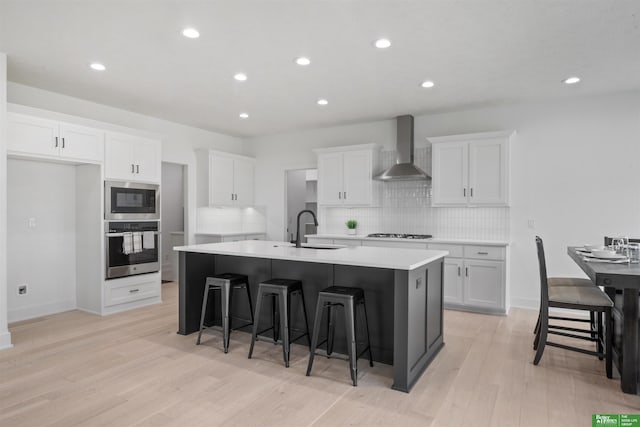 kitchen featuring stainless steel appliances, a sink, white cabinets, wall chimney exhaust hood, and an island with sink