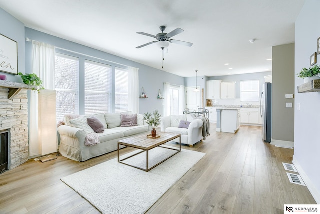 living area featuring light wood-style flooring, baseboards, ceiling fan, and a stone fireplace