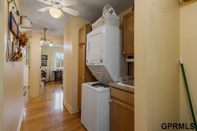 clothes washing area with a textured ceiling, stacked washing maching and dryer, cabinet space, light wood finished floors, and ceiling fan