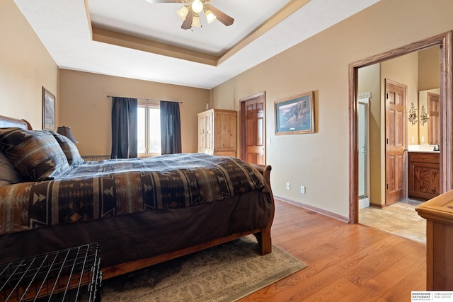 bedroom featuring a raised ceiling, ensuite bathroom, a ceiling fan, light wood-style floors, and baseboards