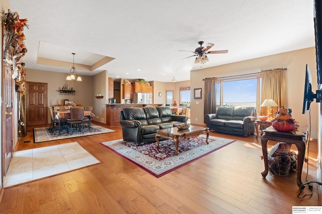 living area featuring light wood-type flooring, a raised ceiling, baseboards, and ceiling fan with notable chandelier