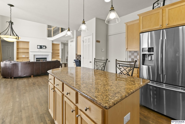 kitchen with a center island, stainless steel fridge with ice dispenser, dark stone counters, a warm lit fireplace, and dark wood-style flooring