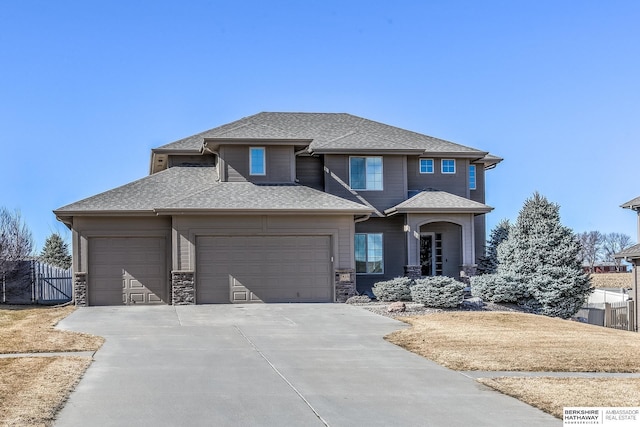 prairie-style home featuring fence, driveway, an attached garage, a shingled roof, and stone siding