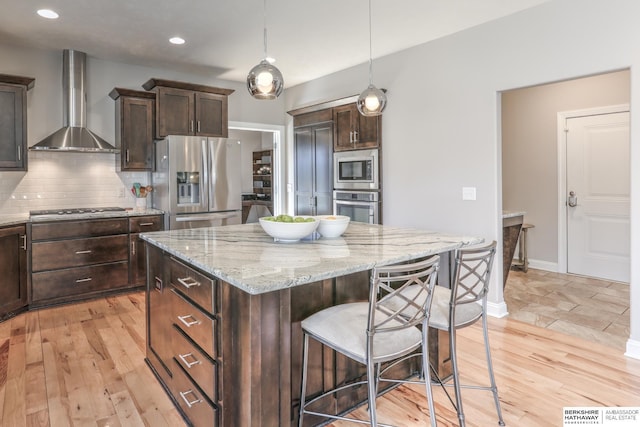 kitchen with light wood finished floors, a breakfast bar, appliances with stainless steel finishes, wall chimney range hood, and tasteful backsplash