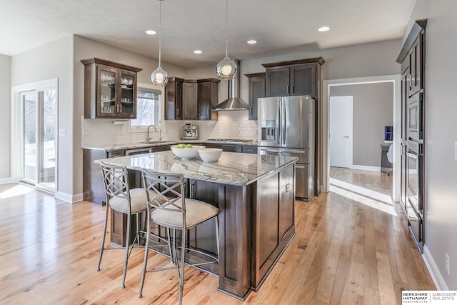 kitchen with stainless steel appliances, light wood-style floors, a breakfast bar area, wall chimney exhaust hood, and light stone countertops