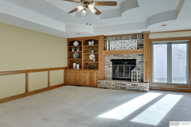 unfurnished living room featuring a brick fireplace, built in shelves, ceiling fan, a tray ceiling, and light carpet