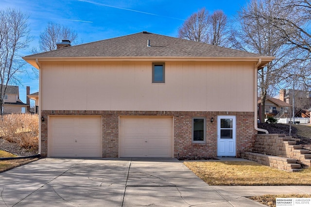 view of home's exterior with concrete driveway, an attached garage, a shingled roof, brick siding, and a chimney