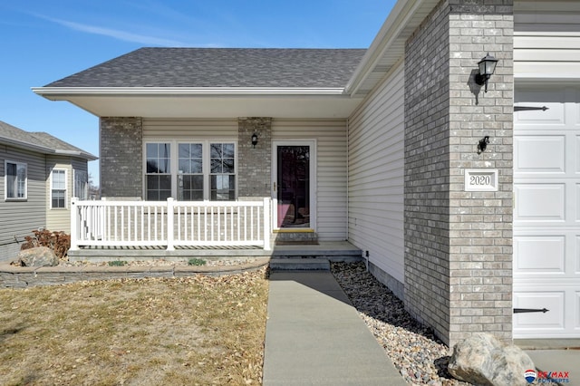 view of exterior entry featuring a garage, brick siding, a porch, and a shingled roof