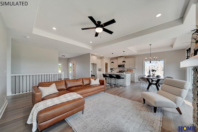 living room with ceiling fan with notable chandelier, a tray ceiling, recessed lighting, and wood finished floors