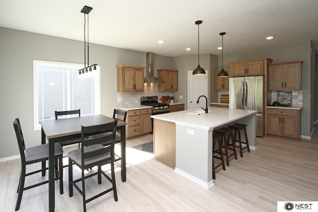 kitchen with a kitchen island with sink, light wood-style flooring, a sink, stainless steel appliances, and wall chimney exhaust hood