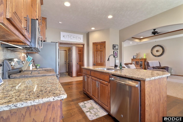 kitchen featuring brown cabinets, an island with sink, a sink, dark wood-style floors, and appliances with stainless steel finishes