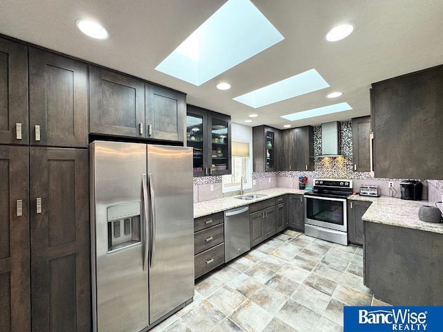 kitchen with a sink, stainless steel appliances, a skylight, wall chimney range hood, and glass insert cabinets