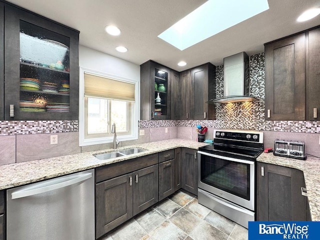 kitchen with a sink, light stone counters, stainless steel appliances, wall chimney exhaust hood, and a skylight