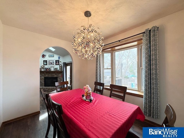 dining area featuring baseboards, arched walkways, dark wood-type flooring, a textured ceiling, and a brick fireplace