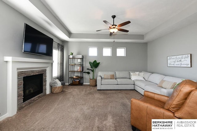 living room featuring carpet, baseboards, a tray ceiling, a fireplace, and ceiling fan